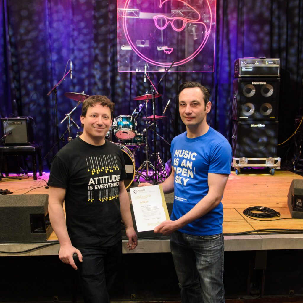 Two white men stand in front of a stage in a venue. One of them is holding up a certificate.