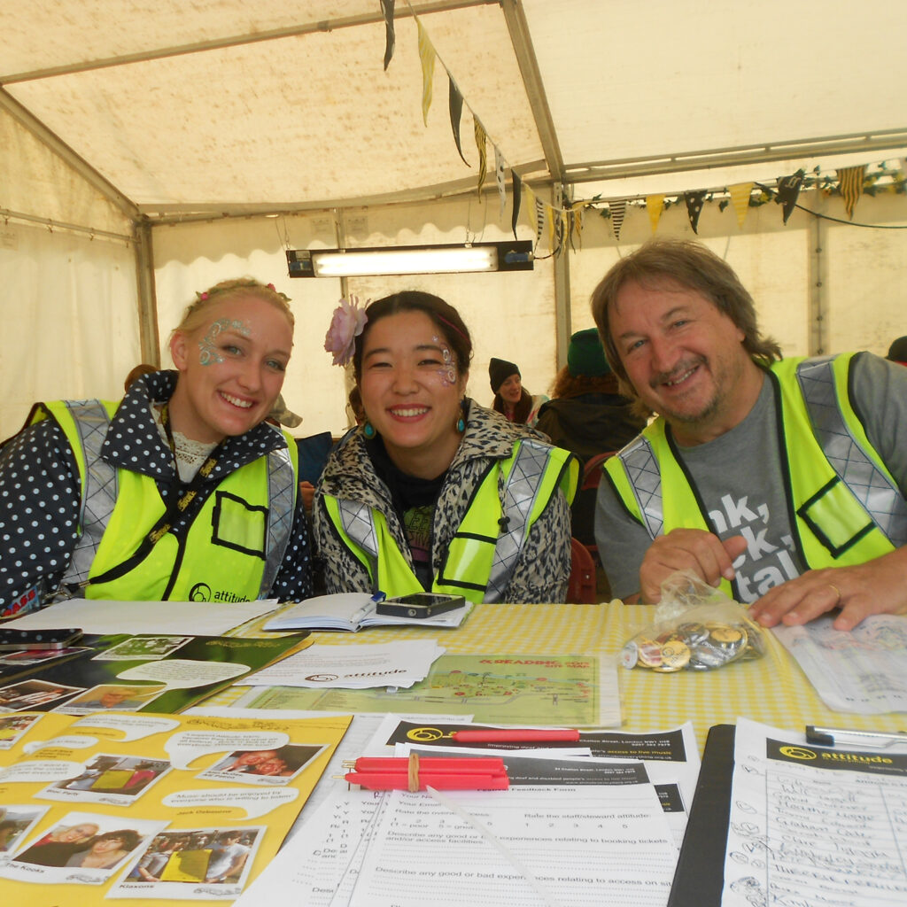 Three volunteers in high vis jackets sat behind a desk in a festival information tent. 