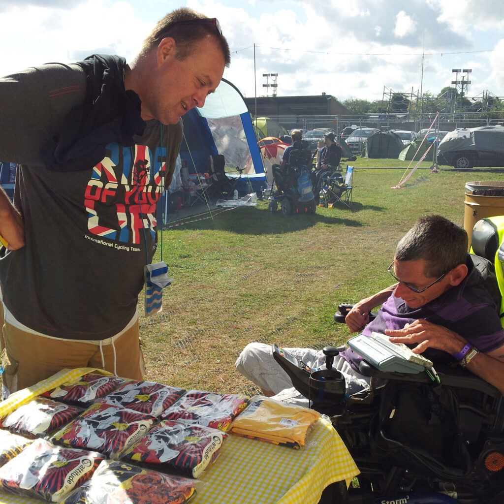 A man leaves over a table covered in T-shirts whilst another man types on a communication device on his power wheelchair.