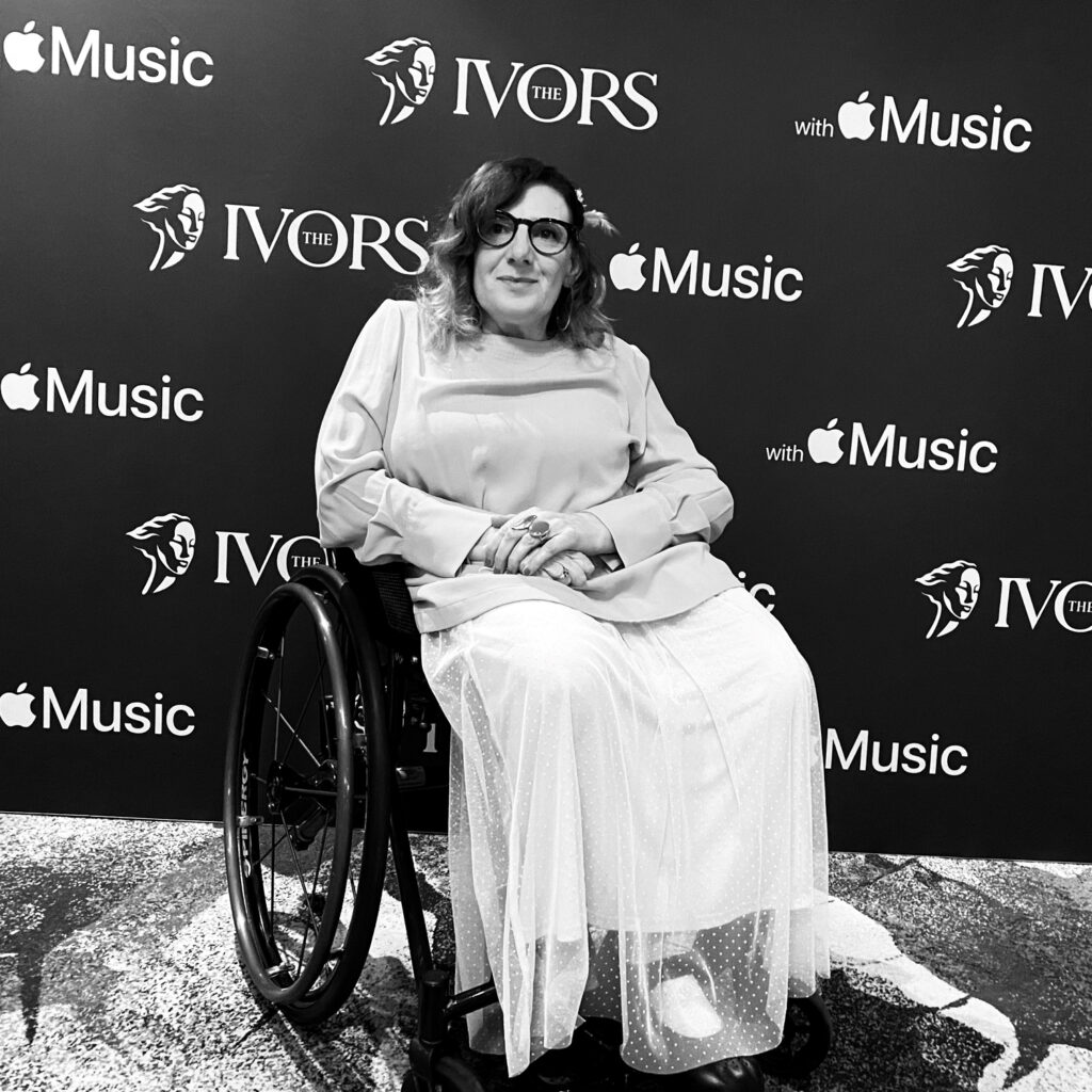 A blonde woman wearing glasses, a long sleeve top and lacy skirt sits in a wheelchair in front of a wall with the words The Ivors printed on it