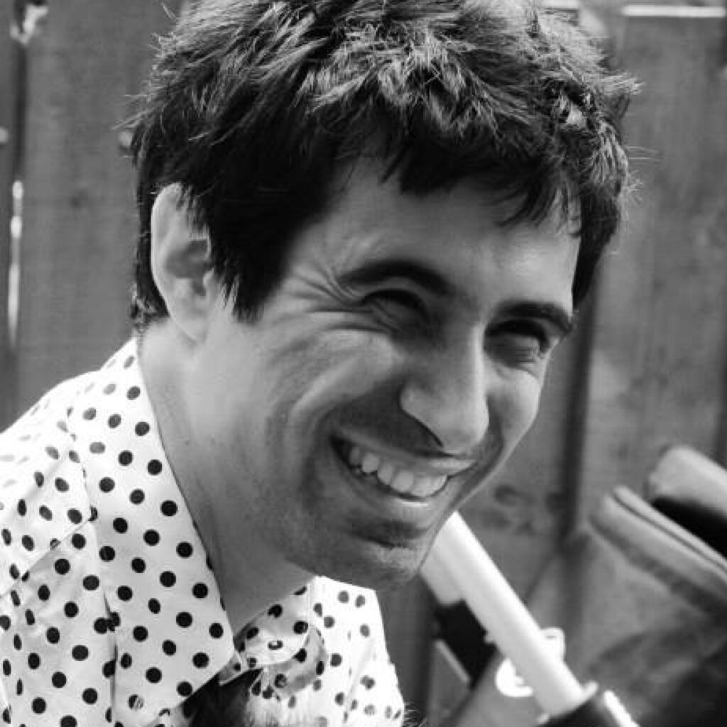 A man with short black hair and stubble in a spotted shirt laughing.