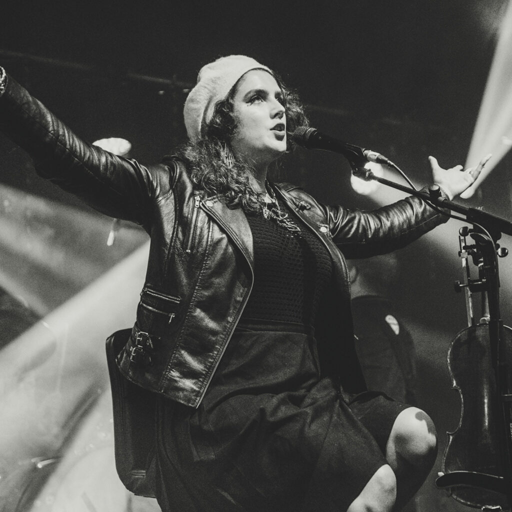 A white woman with long dark hair wearing a leather jacket and raising her arms on stage.