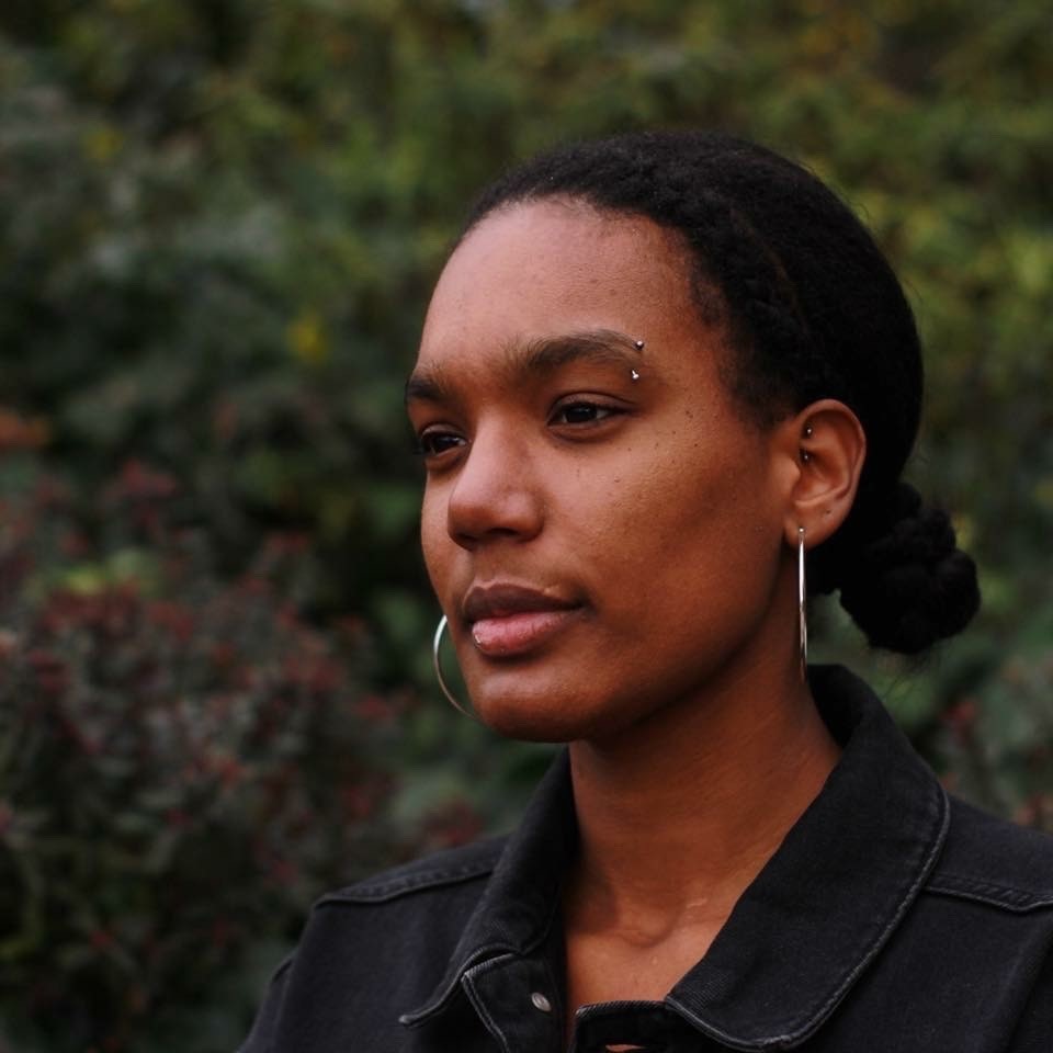 A young black woman with tied back black hair and hoop earings standing in from of green leaves