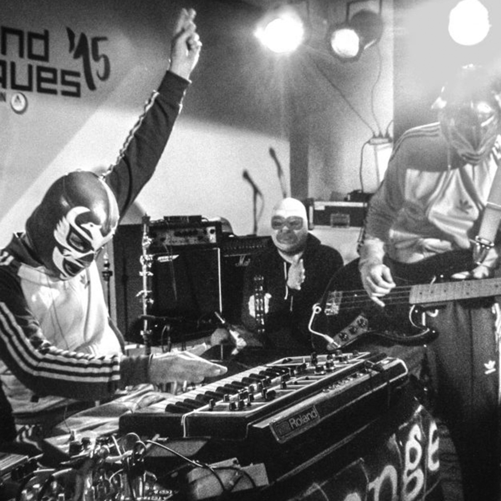 A band performing on stage wearing wrestling masks. The keyboardist is seated with a hand in the air.