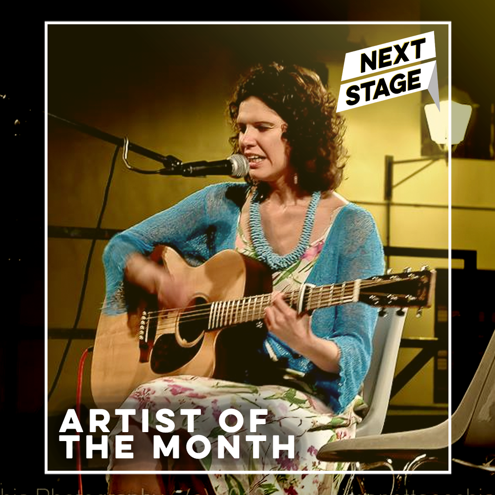 image of Elena Piras sitting on chair playing an acoustic guitar and singing, she is wearing a blue cardigan and a colourful dress. The image has the text 'Artist of The Month' on it and the Next Stage logo.