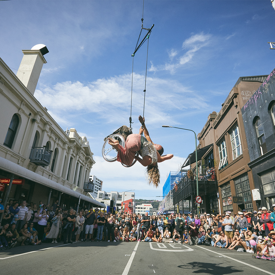 A person using a wheelchair hangs from a trapesze above a street, watched by a crowd of people.