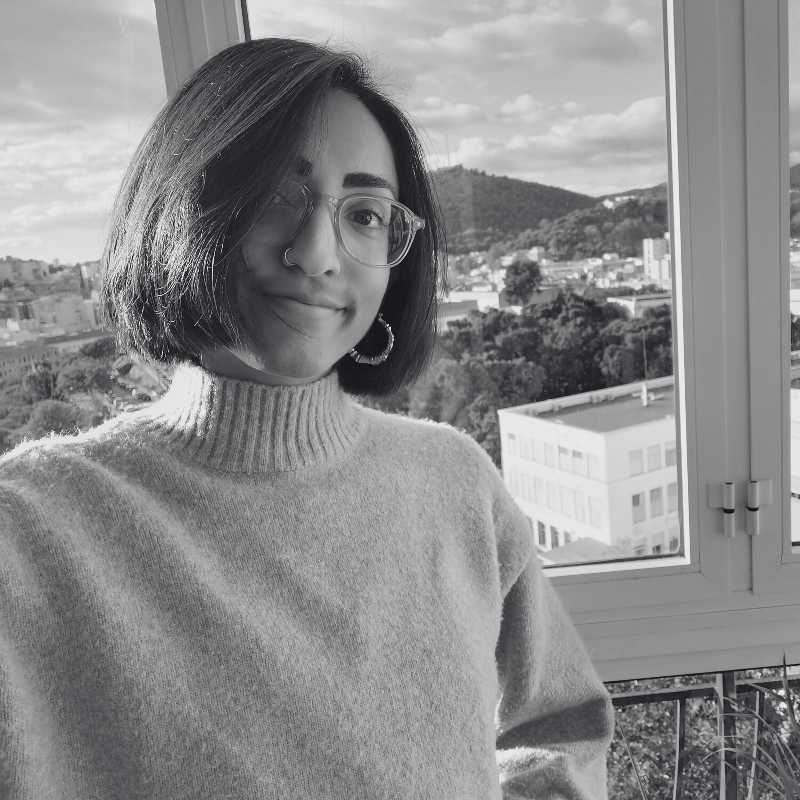 Henna wears a turtleneck jumper, gold hoops, nose ring and goggle like glasses. They’re standing on a balcony in Malaga with buildings and lush mountains behind. They’re smiling and looking into the camera.