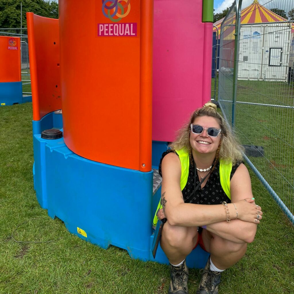 Fern Beard crouching by PEEQUAL festival loos. Fern is a white feminine person, blonde hair half tied up wearing sunglasses and a hi-vis.