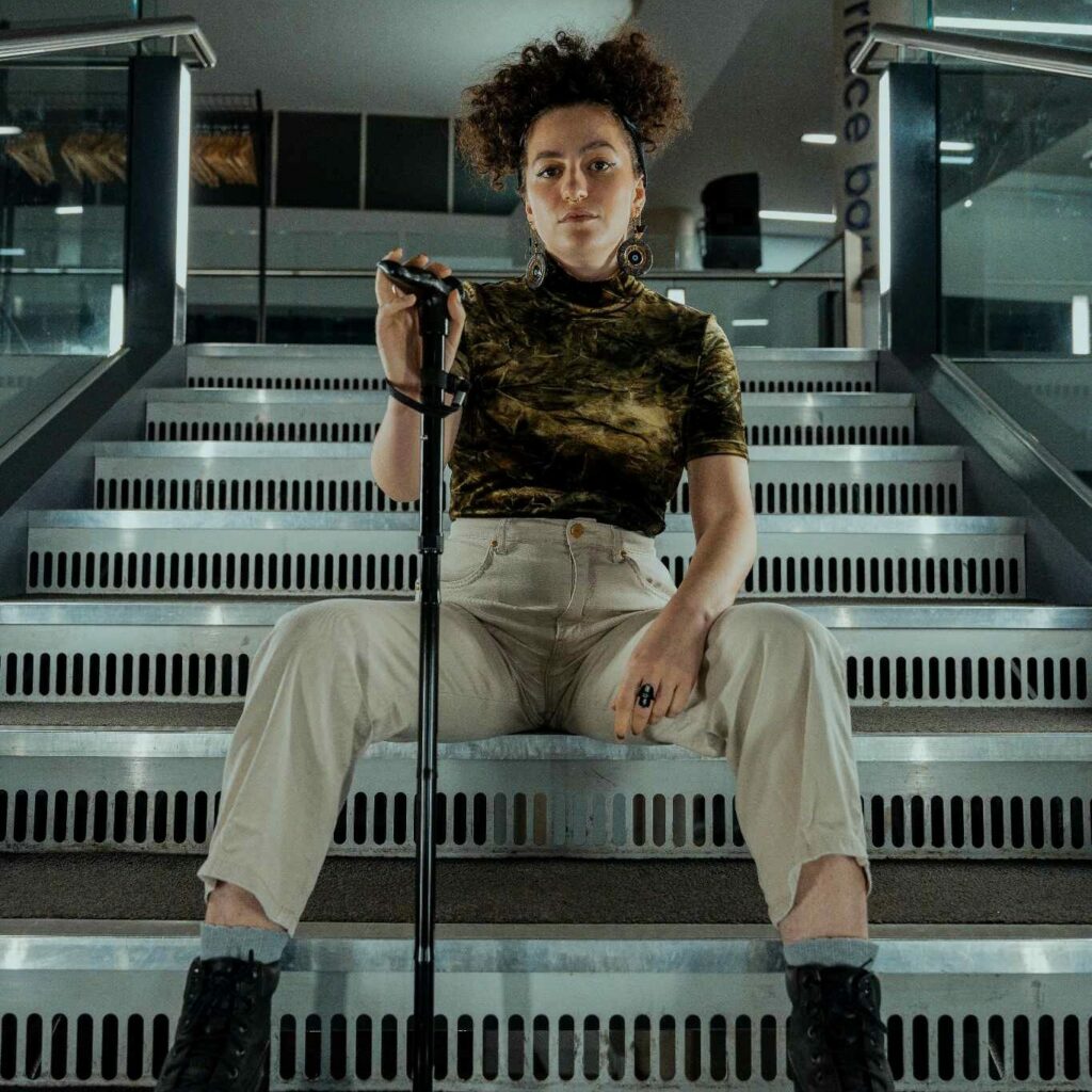 Jess Ansell sitting on a staircase staring into the camera holding her walking stick. Jess is a racially ambiguous feminine person with dark curly hair tied up.