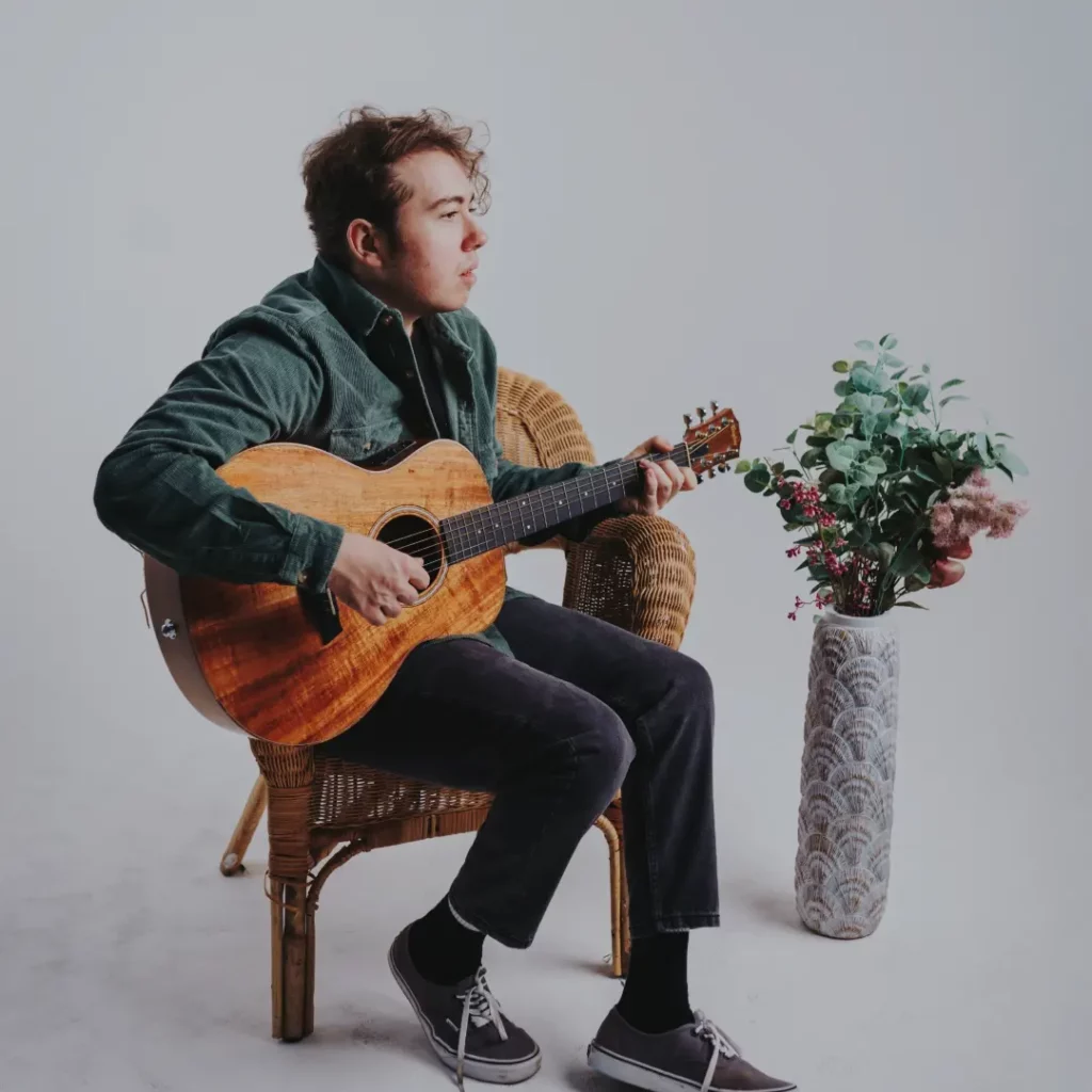 Louis sitting on wicker armchair strumming acoustic guitar. he wears a jacket, trousers, and Vans shoes. There's a tall vase with flowers on the floor beside him