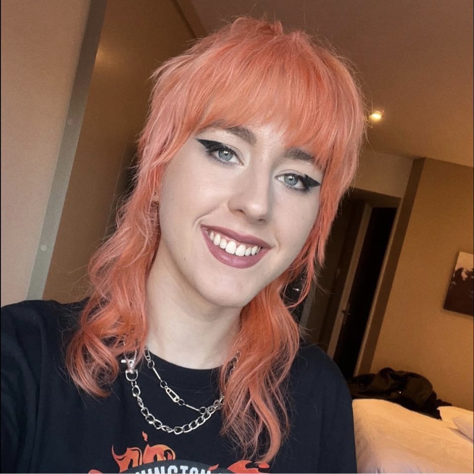 Headshot of Taylor Booth smiling into camera, midlength curly hair & full fringe, black winged eyeliner, lipstick, wearing tshirt & silver chains