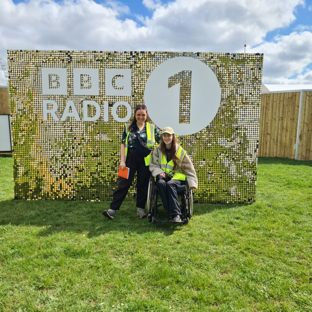 Alex Covell and Sakara at BBC Radio 1 Big Weekend Luton. They are in front of a big glittery, gold sign reading BBC Radio 1. They are both white women with long brown hair wearing yellow high vis vests. Sakara is a wheelchair user.