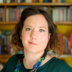 Headshot of Kate Carmichael looking into the camera, in front of a blurred bookshelf. Kate is a white feminine person with short brunette hair, wearing a blue and green necklace and a green top.