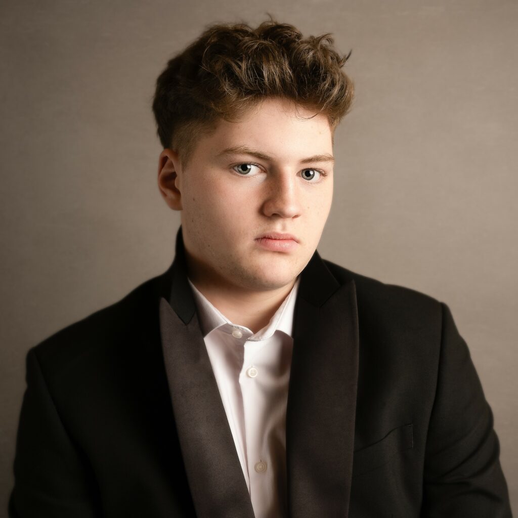 Headshot of Finn Simpson-Murphy looking seriously, against a neutral-tone background. Finn is a white person with short brown hair wearing a black tuxedo jacket with satin lapels over a white shirt.