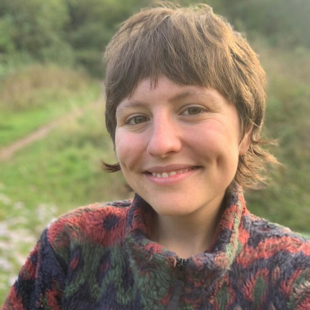 Georgina Daniels enjoying the outdoors smiling. Georgina is a white feminine person with short brunette hair wearing a thick, textured fleece.