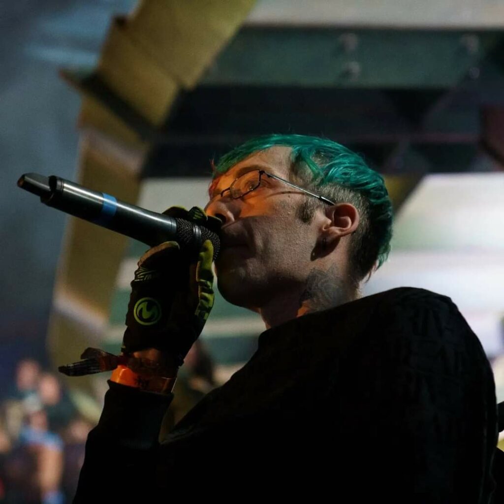 Mark Humphries rapping into a microphone at a performance. Mark has teal short hair wearing all black, and some green details on his gloves. He wears glasses and has tattoos on his neck.