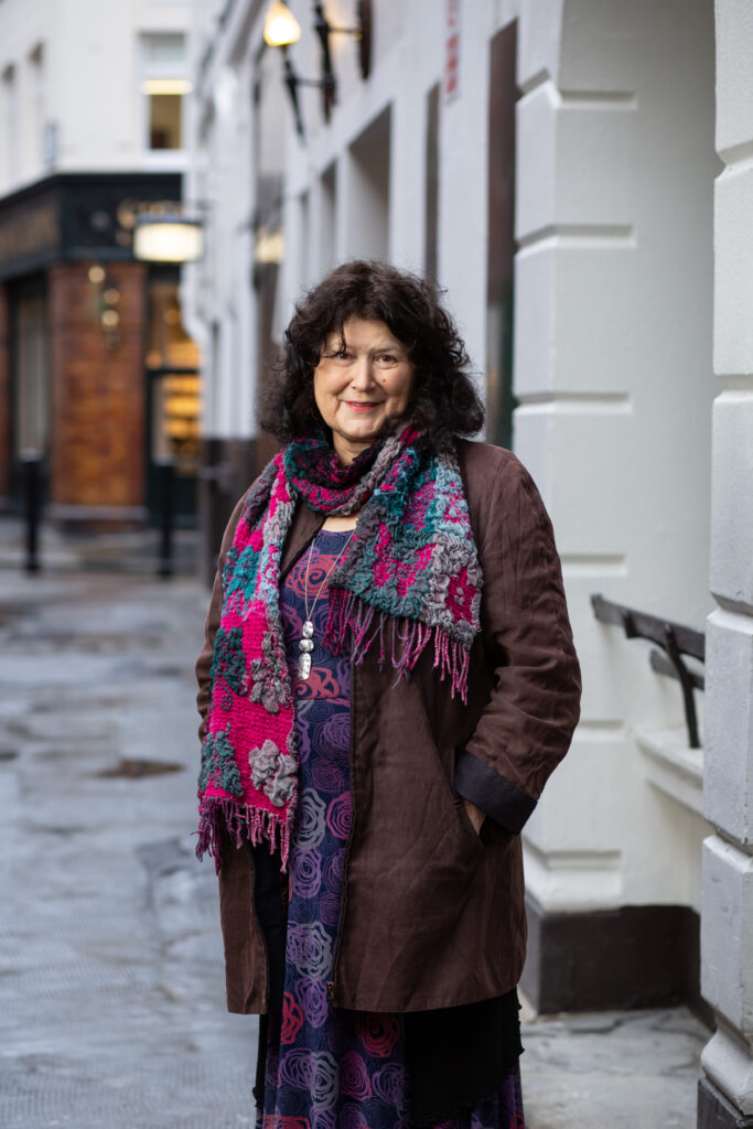 Claire Cordeaux, a woman with brown hair, is standing with her hands in her pockets. She is wearing a long brown coat and a colourful scarf.