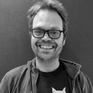 A black and white headshot of Paul, a white man with short hair and wearing glasses, smiling at the camera.