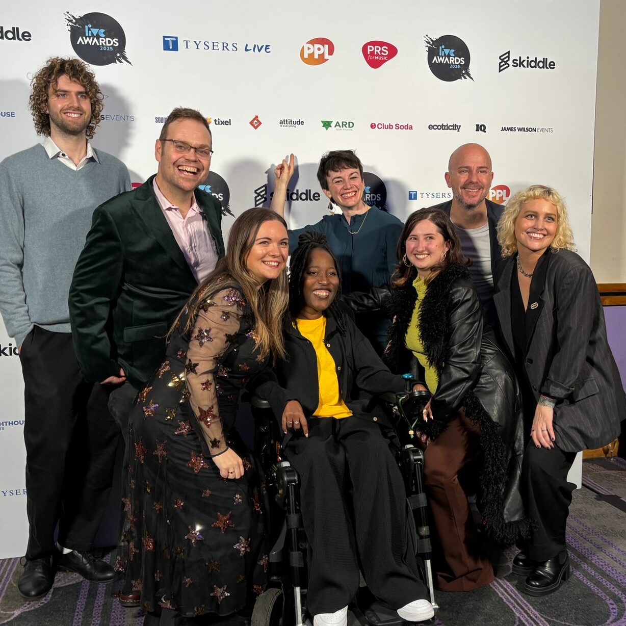 Eight staff members from Attitude is Everything, Johann, Paul, Catherine, Andrew, Rhiannon, Jessica, Jacqui and Alex, huddled together in smart casual wear, smiling in front of a board of partner and sponsor logos from the LIVE Awards 2025.