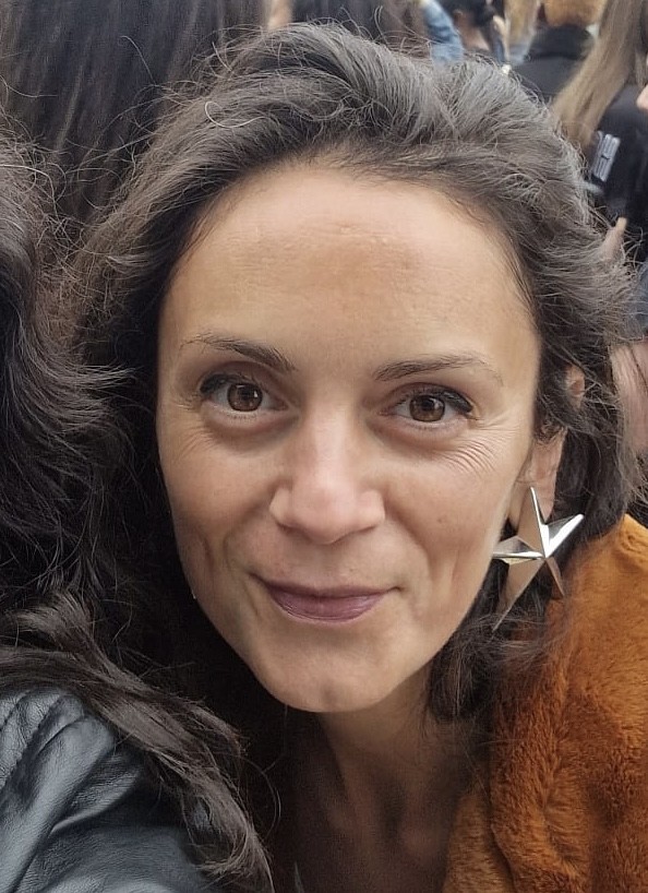 A close-up of Lish, a white woman with dark brown hair looking at the camera. She's wearing large star-shaped silver earrings. She is happy as she is at a gig; in the background, there are other gig-goers in the distance. 