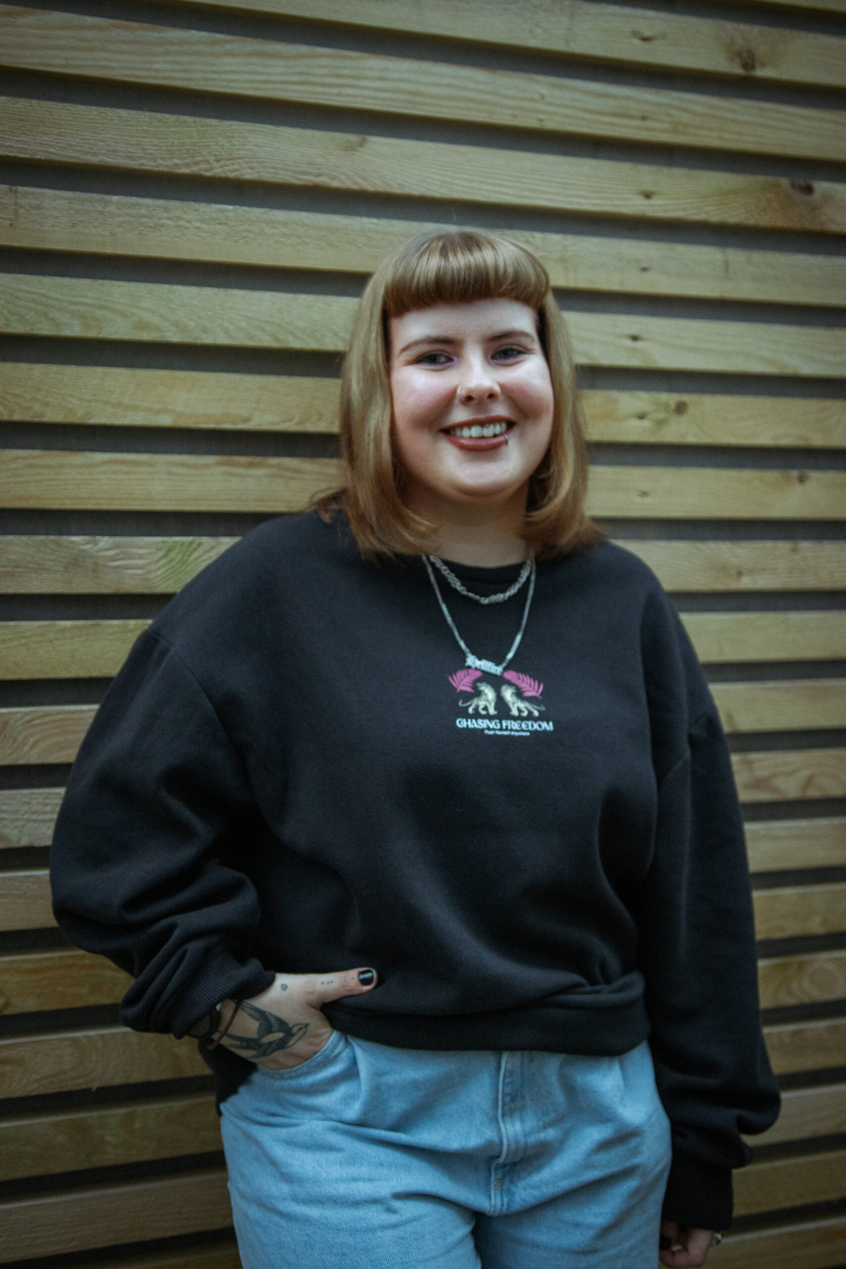 Bee, a white femme person with mousy brown shoulder length hair and a fringe, is looking directly at the camera with a wide smile. They are wearing a black jumper and silver layered necklaces the jumper has two tigers on with the words ‘chasing freedom’. There is fencing in the background of the picture.   