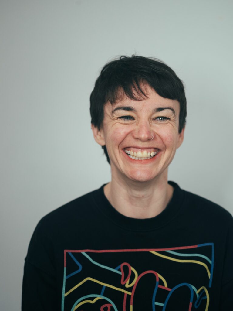 Catherine, a white, femme person with short, dark hair is smiling. They are wearing a black jumper with a sloth on it and have a nose piercing. 