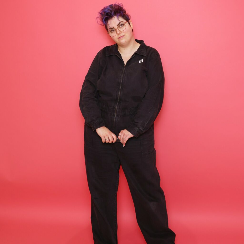 A white person with short purple hair and glasses stands against a bright pink backdrop. They wear a black jumpsuit and look at the camera with a relaxed, slightly tilted posture, holding their hands loosely together in front.