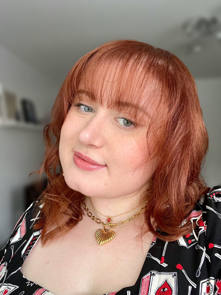 Maia, a white femme person with auburn hair, is smiling at the camera with their head tilted to the side. They are wearing a black, red and white patterned shirt and layered gold necklaces.
