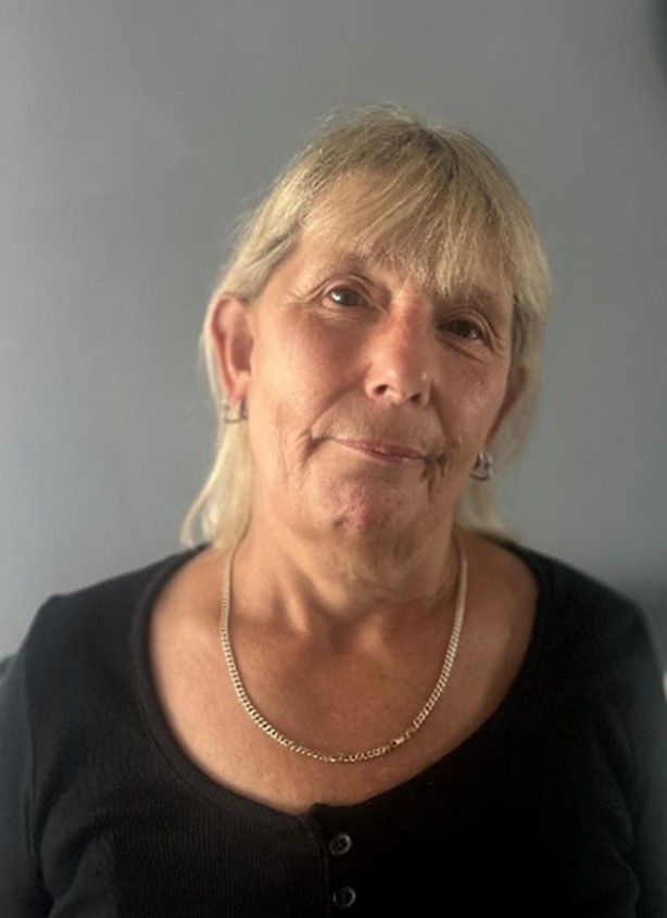 Sarah, a white middle-aged woman with blond hair, smiling at the camera and wearing a black top and a gold necklace.