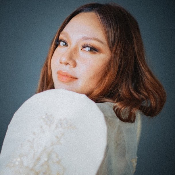 A portrait of Sha aka SO SHA, a Filipina artist, looking towards the camera with a soft, calm expression. She wears a cream Filipiniana-inspired embroidered sleeve and has shoulder-length auburn hair. The background is a smooth teal-grey, and the lighting is gentle and warm.