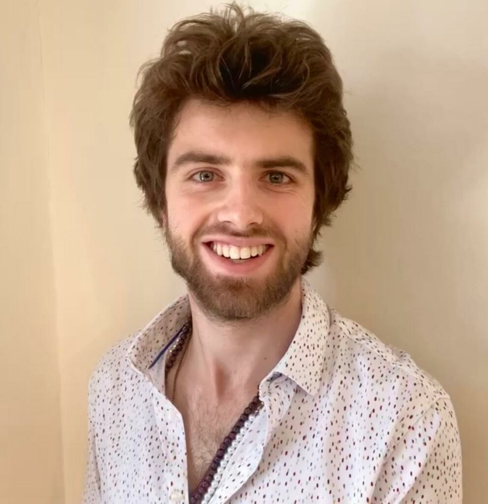 Johann, a young man with thick, slightly tousled brown hair and a short beard smiles at the camera. He is wearing a white shirt with small multicoloured dots, and stands against a plain light-coloured wall.