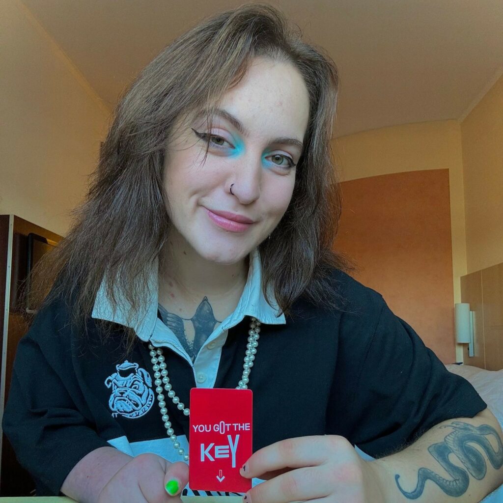A white woman with shoulder-length brown hair and teal eye makeup smiles at the camera. She wears a black collared shirt with a pearl necklace and has tattoos on her arm. Keys holds up a red card that says “YOU GOT THE KEY” in white text. The photo is taken indoors with warm lighting. 