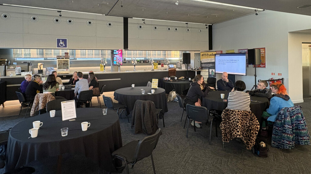 Beside a café and bar are some big tables and chairs, with two tables full of people from the Bristol Accessibility Afternoon discussing access. In the corner of the room, a PowerPoint presentation and an Attitude is Everything banner beside. 