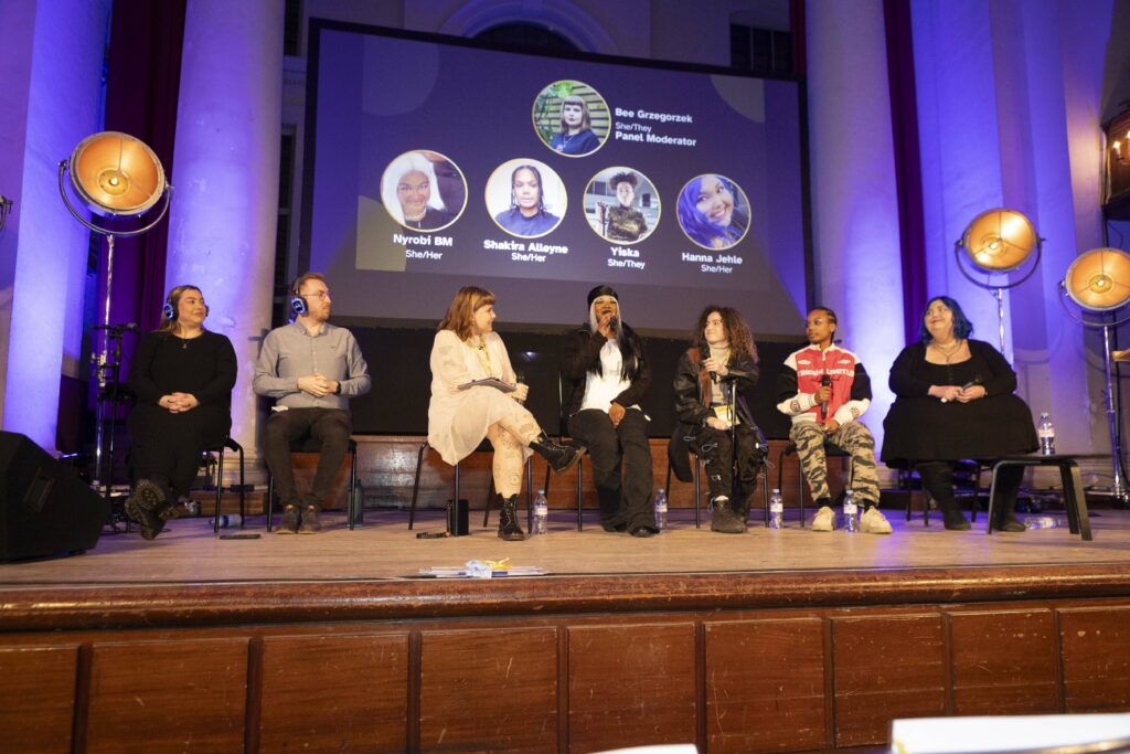 Seven people are sat on a stage as part of a panel session. They are holding microphones. Two people on the far left of the stage are British Sign Language interpreters and are wearing large blue headphones.