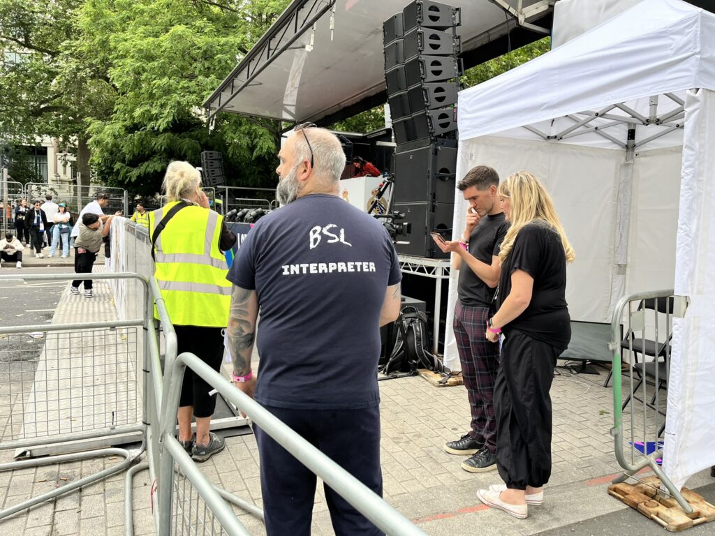A British Sign Language interpreter is standing with their back facing the camera, facing a stage. Their t shirt has writing on the back to identify that they are a BSL interpreter. 