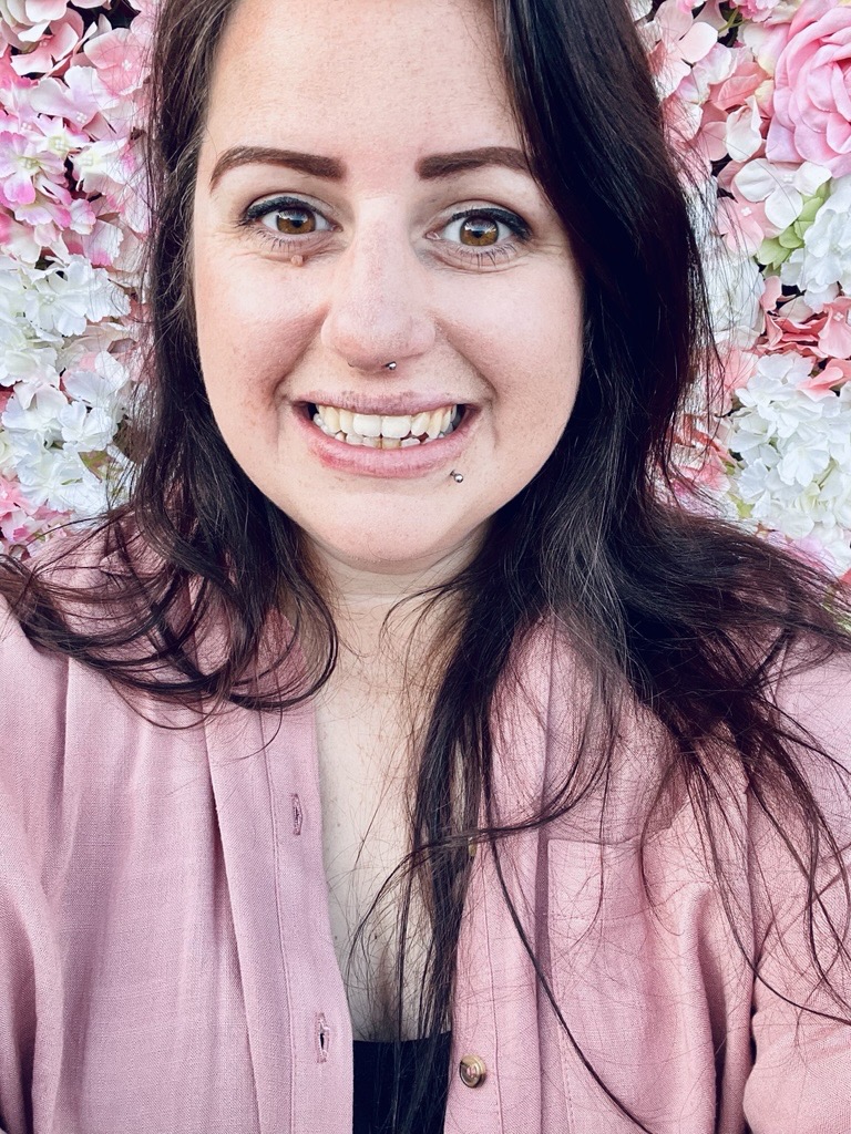 Becki Wingell, a white woman with brown hair smiling widely. She's wearing a pink shirt, standing in front of a wall of pink and white flowers.
